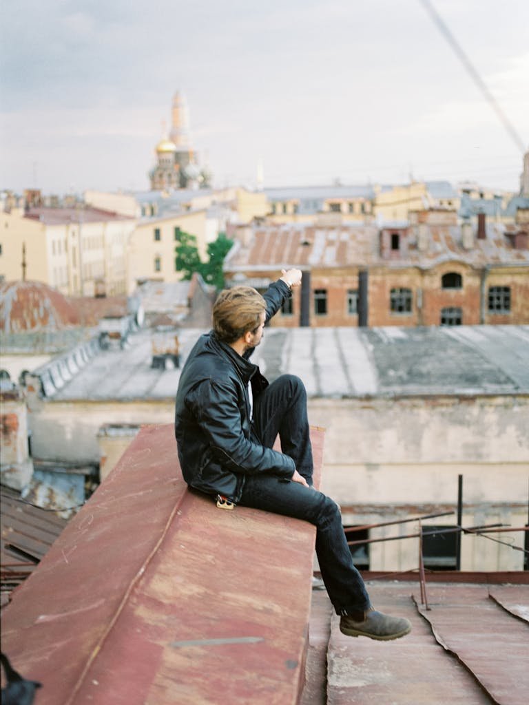 Man in jacket sitting on a rooftop in urban area, with city buildings in background, pointing at the horizon.