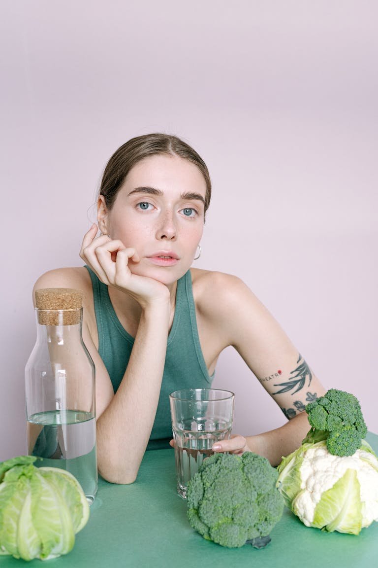 Woman sitting with a glass of water surrounded by fresh vegetables including broccoli and cabbage.